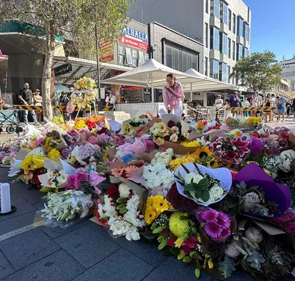 A large memorial with colorful flower bouquets on a city street, surrounded by people observing respectfully.